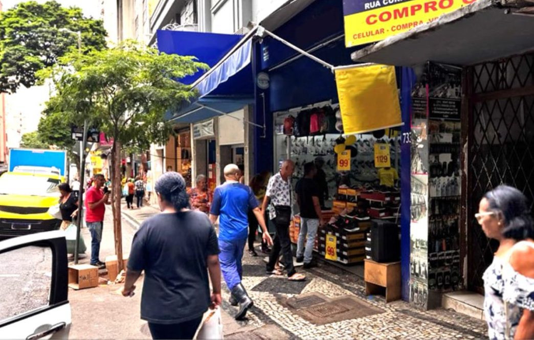 Pessoas caminhando em calçada movimentada com lojas.