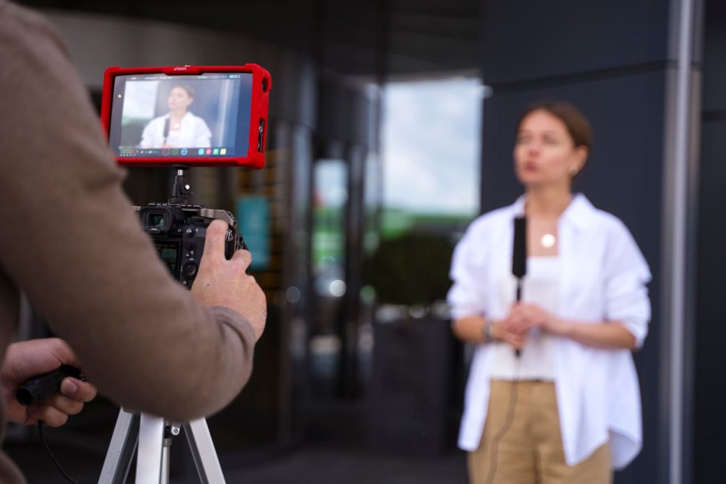 Profissional de multimídia sendo gravada durante entrevista externa, com cinegrafista operando câmera e monitor de vídeo.