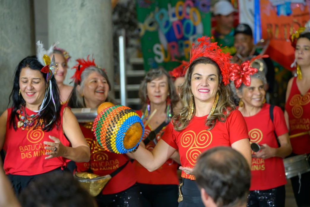Mulheres integrantes de bloco carnavalesco se apresentam com instrumentos de percussão durante o Carnaval nas Cidades Históricas de Minas Gerais, usando camisetas vermelhas e adereços coloridos, em ambiente festivo e cultural.