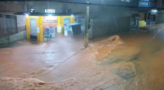 Rua tomada por enxurrada durante temporal em Ubá (MG), com água barrenta invadindo via e estabelecimentos comerciais à noite.