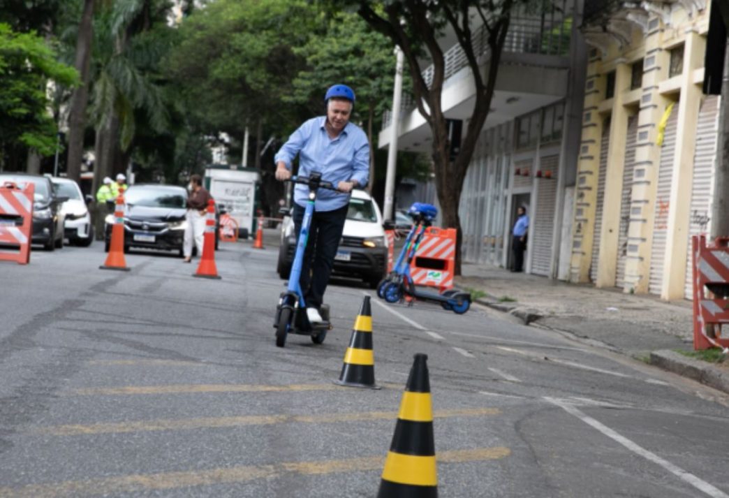 Homem utilizando patinete elétrico em via urbana de Belo Horizonte durante demonstração prática, com cones de sinalização e veículos ao fundo