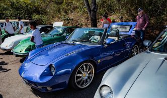 Carros esportivos e clássicos expostos ao ar livre, com destaque para um conversível azul em primeiro plano, enquanto visitantes observam os veículos em um encontro automotivo.