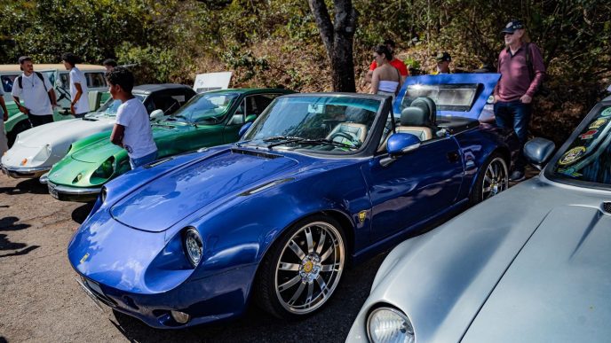 Carros esportivos e clássicos expostos ao ar livre, com destaque para um conversível azul em primeiro plano, enquanto visitantes observam os veículos em um encontro automotivo.
