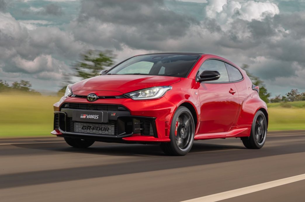 Toyota GR Yaris vermelho em movimento, com design esportivo agressivo, grade frontal ampla, rodas escuras e cenário de estrada ao fundo.