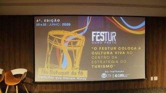 Tela de projeção em auditório exibe a identidade visual do FESTUR Ouro Preto 2026, com ilustração de tambor, datas do evento (10 a 12 de junho de 2026) e o tema “No Batuque da Fé – tradições que movem Minas”, ao lado de uma cadeira em ambiente institucional.