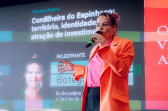 Bárbara Botega fala ao microfone durante palestra, diante de telão sobre território, turismo e atração de investimentos.