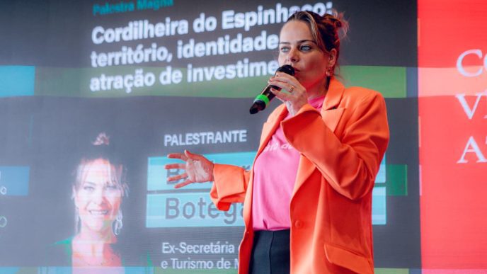 Bárbara Botega fala ao microfone durante palestra, diante de telão sobre território, turismo e atração de investimentos.