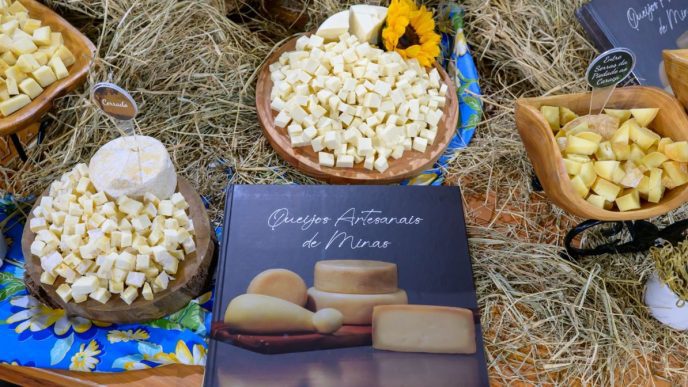 Mesa de exposição com Queijo Minas Artesanal em cubos, peças inteiras, palha decorativa e livro sobre queijos artesanais de Minas.