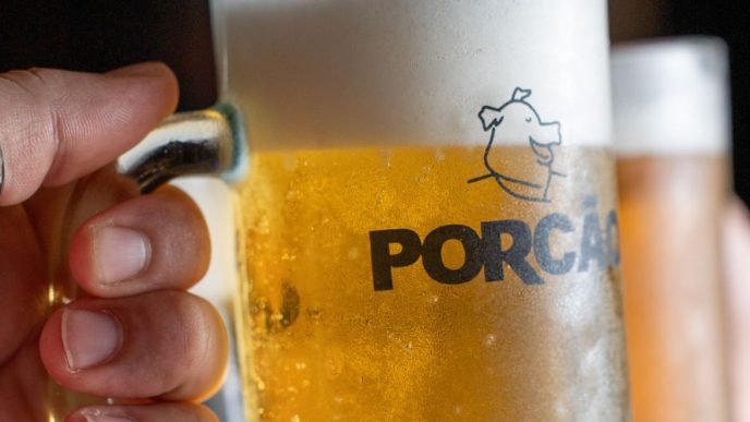 Caneca de chope gelado do Porcão BH em primeiro plano, segurada por uma pessoa, com outra bebida desfocada ao fundo.