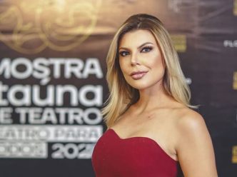 Mulher loira usando vestido vermelho posa em frente a painel de evento, com expressão confiante, representando matéria sobre comunicação e oratória em Belo Horizonte.