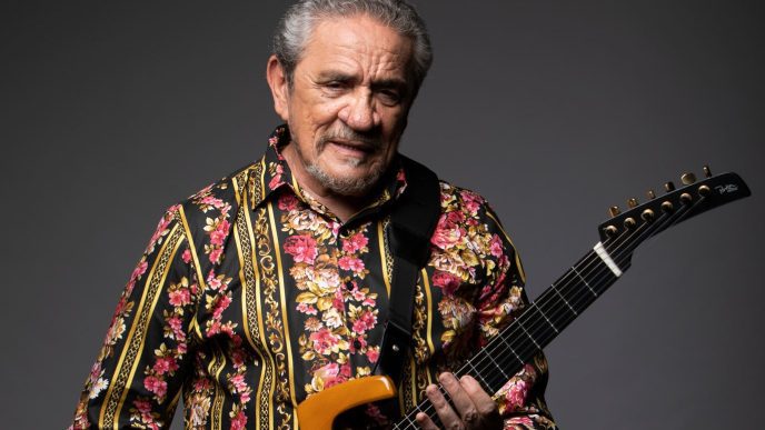 Zé Ramalho posa diante de um fundo escuro, segurando uma guitarra preta e usando uma camisa estampada com flores e detalhes dourados.