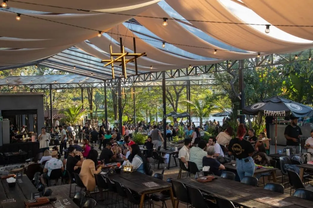Ambiente amplo e coberto do Bebedouro Bar, decorado com iluminação suspensa e teto translúcido, repleto de mesas ocupadas por pessoas confraternizando durante o evento da Churrascada Julina, com vegetação ao fundo e clima descontraído.