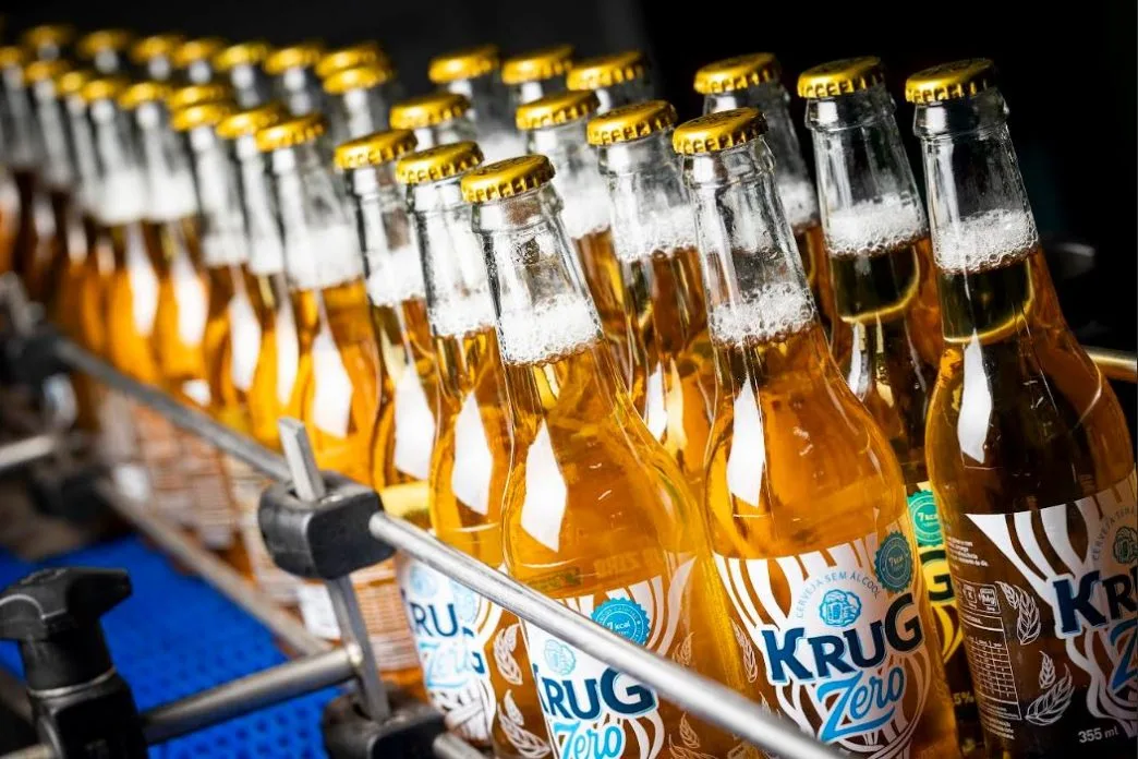 Garrafas de Krug Zero organizadas em linha de produção industrial, com rótulos visíveis e líquido dourado brilhante, destacando a nova cerveja sem álcool da marca mineira Krug Bier.