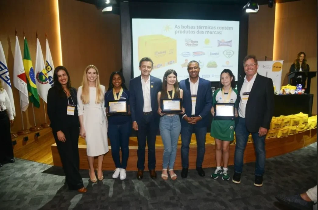 Grupo de estudantes premiados, acompanhados por organizadores e representantes do SILEMG, posam sorridentes em cerimônia de premiação do Concurso SILEMG de Desenho e Redação 2025. Ao fundo, há um telão com marcas apoiadoras e bolsas térmicas amarelas com produtos lácteos.