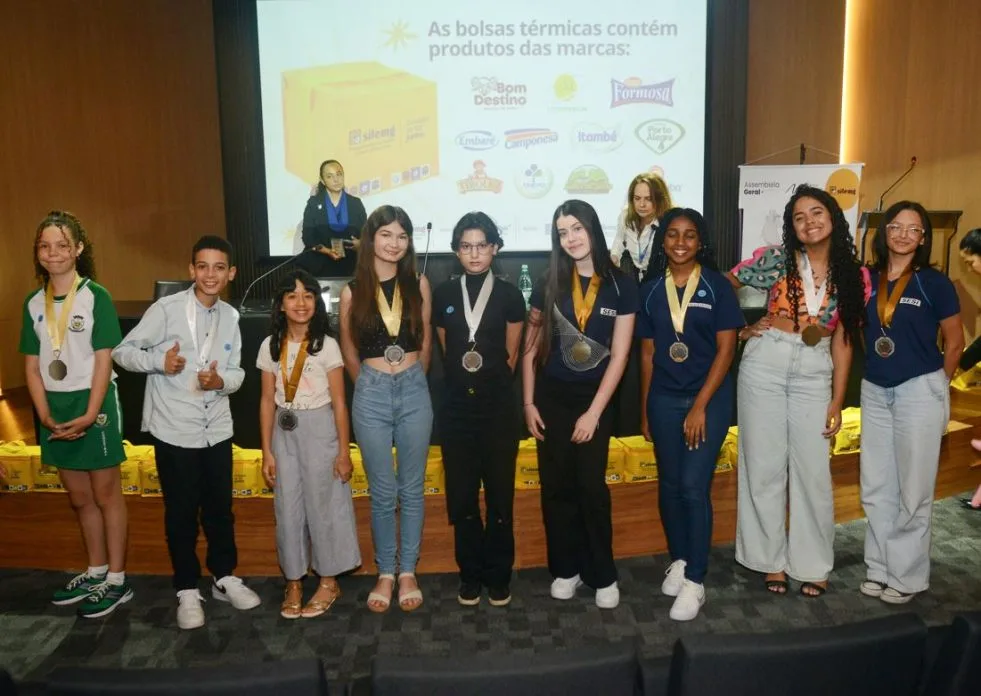 Estudantes premiados com medalhas posam sorridentes no palco durante a cerimônia do Concurso SILEMG de Desenho e Redação, edição 2025, realizada em Belo Horizonte. Ao fundo, painel destaca marcas parceiras e bolsas térmicas com produtos lácteos.