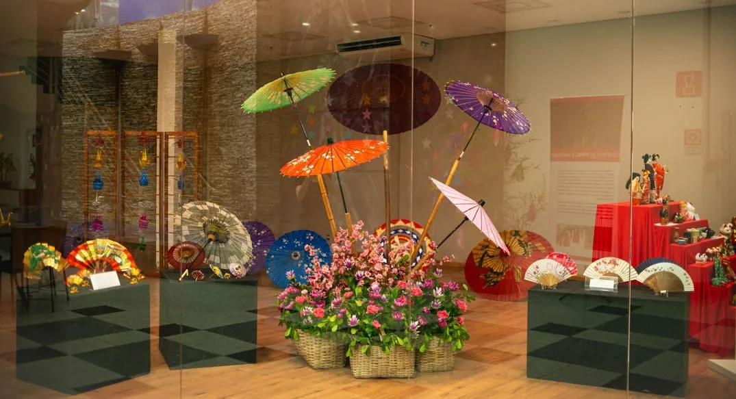Exposição cultural japonesa com guarda-chuvas tradicionais coloridos, leques decorativos e arranjo floral, instalada em espaço interno com vitrines e objetos típicos do Japão no Festival Tanabata Matsuri em Belo Horizonte.