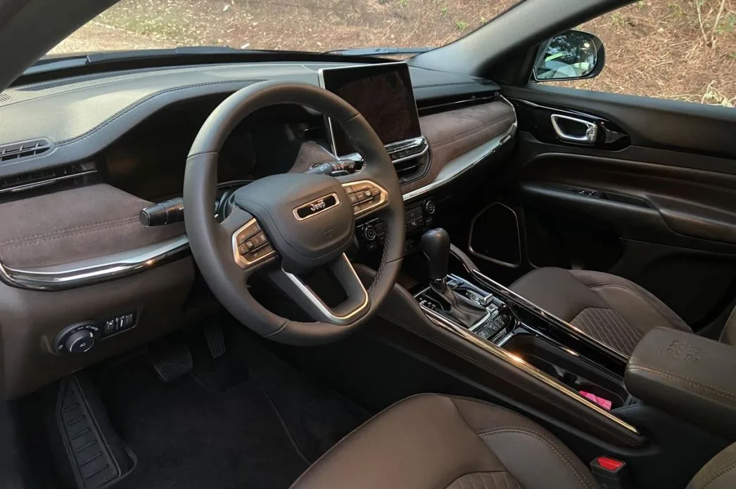 Interior do Jeep Commander Overland 2025 com acabamento refinado em couro e suede, volante multifuncional com comandos integrados, central multimídia flutuante, câmbio automático e detalhes cromados no console e nas portas.