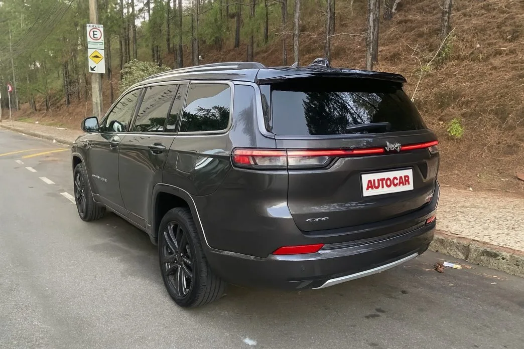Vista traseira do Jeep Commander Overland 2025 estacionado em rua arborizada, destacando as lanternas em LED com assinatura horizontal contínua, rodas esportivas, vidros escurecidos e a placa personalizada da Autocar aplicada para avaliação editorial.