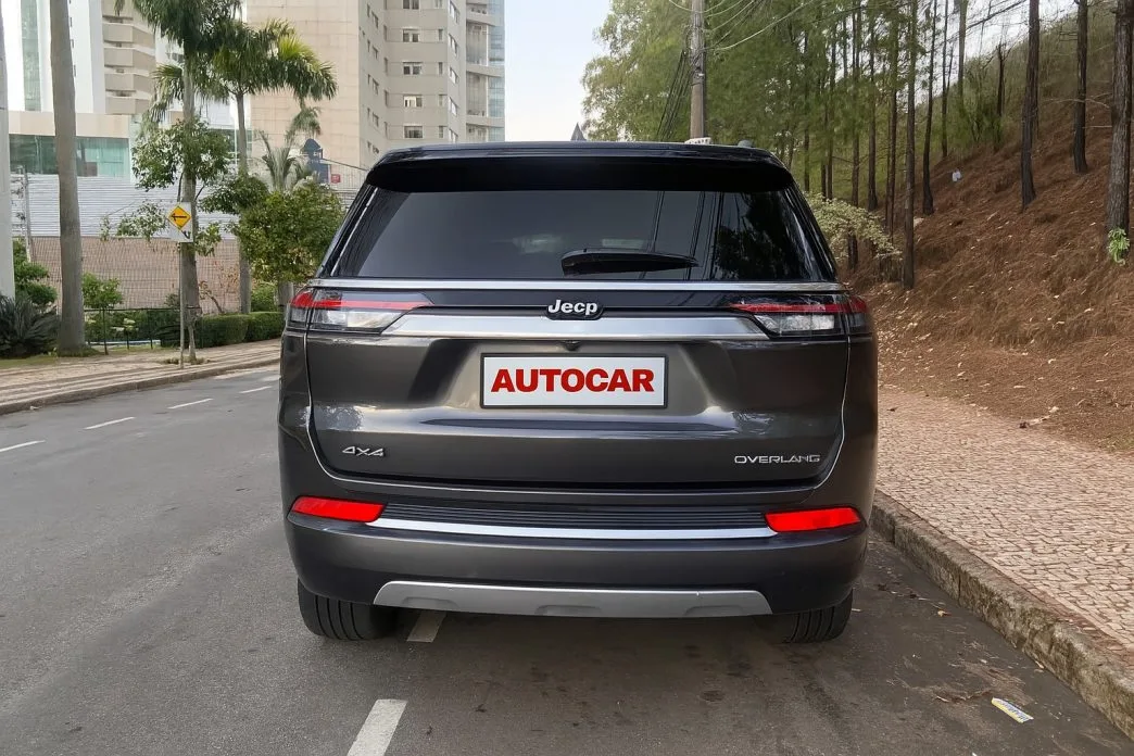 Traseira do Jeep Commander Overland 2025 4x4 estacionado em via urbana com paisagem mista de prédios e árvores, destacando a barra luminosa em LED, logotipo Jeep centralizado e a placa personalizada Autocar.