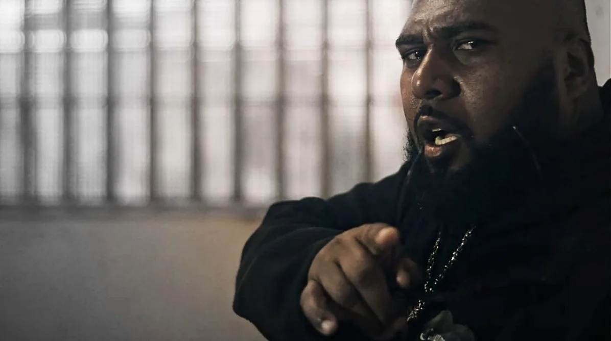 Black W em cena impactante do clipe “Lágrimas de Um Detento” — com expressão firme e dedo apontado para a câmera, o rapper mineiro transmite urgência e confronto direto com o sistema opressor retratado na música. Ao fundo, grades desfocadas reforçam a atmosfera carcerária, enquanto sua presença poderosa encarna a voz dos silenciados.