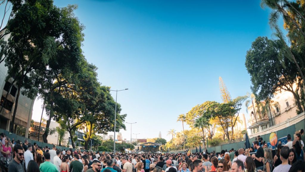 Italianíssima transforma BH em reduto da cultura italiana 4 Multidão reunida em uma rua arborizada de Belo Horizonte durante um evento cultural ao ar livre, sob céu azul e clima ensolarado, com um palco ao fundo e pessoas interagindo alegremente.