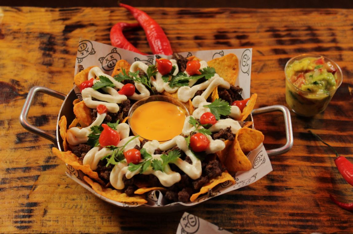 Prato de nachos servido em panela metálica, com carne moída apimentada, sour cream, cheddar, pimentas e coentro fresco, acompanhado de guacamole em copo, sobre mesa rústica de madeira.
