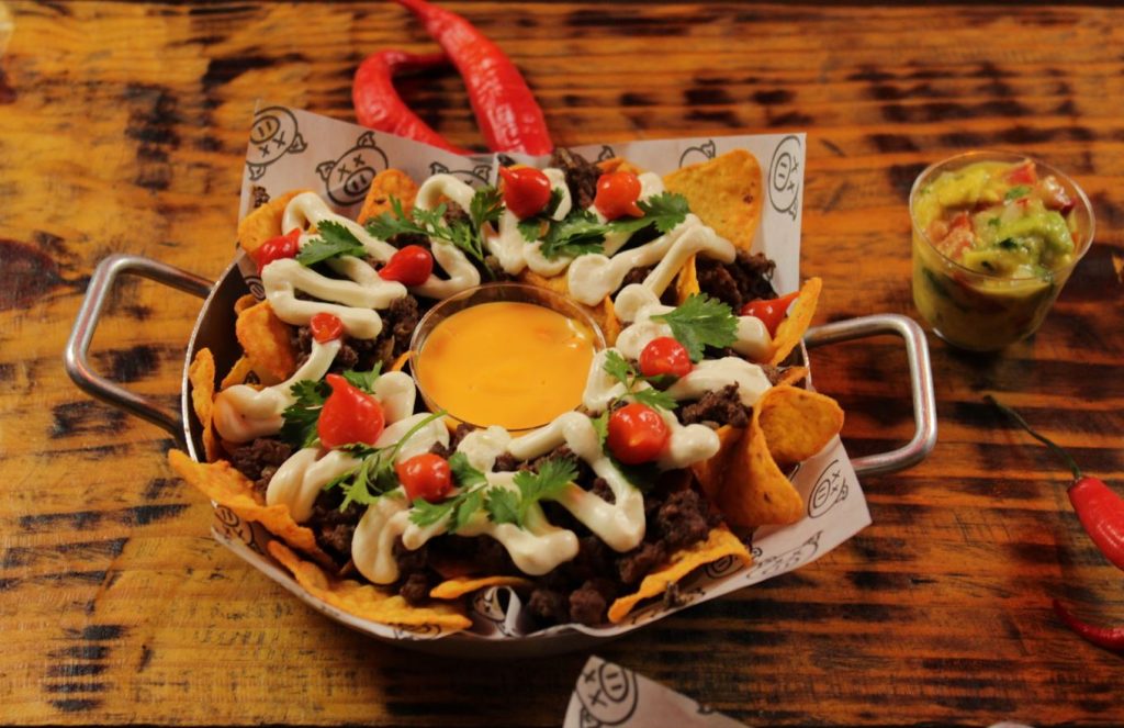 Prato de nachos servido em panela metálica, com carne moída apimentada, sour cream, cheddar, pimentas e coentro fresco, acompanhado de guacamole em copo, sobre mesa rústica de madeira.