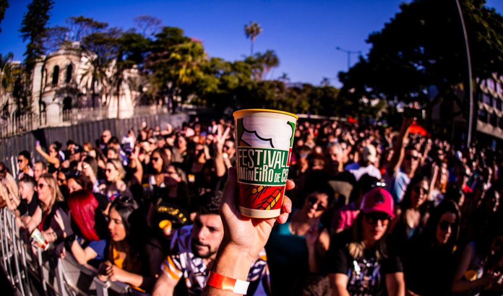 Mão segurando um copo do Festival Mineiro da Cerveja em meio a uma multidão animada sob céu ensolarado, durante apresentação ao ar livre.