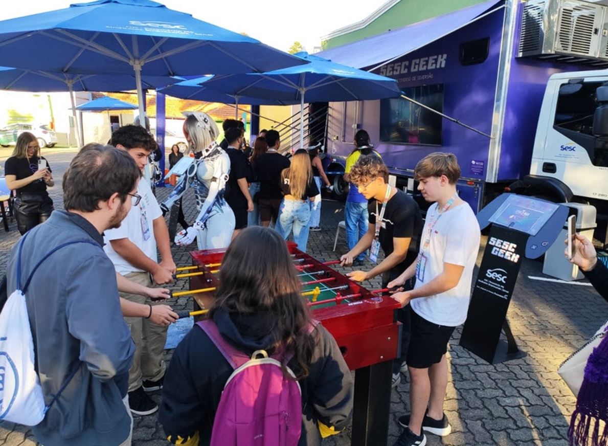 Jovens interagem em uma partida de totó em espaço ao ar livre do Sesc Geek, durante o HackTown 2025, com presença de cosplayer robótica futurista e estrutura móvel do Sesc ao fundo.