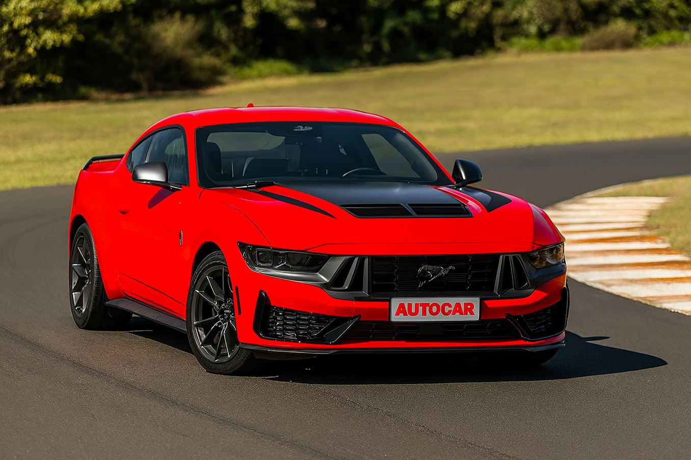Fotografia de um Ford Mustang Dark Horse vermelho, com detalhes em preto no capô e na grade frontal, em movimento em uma pista de asfalto em curva, durante o dia. A placa frontal exibe a logomarca “Autocar”.