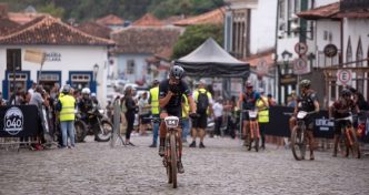 Ciclista cruza a linha de chegada na Praça Gomes Freire, em Mariana (MG), durante o Iron Biker Brasil 2025, cercado por público e construções históricas.