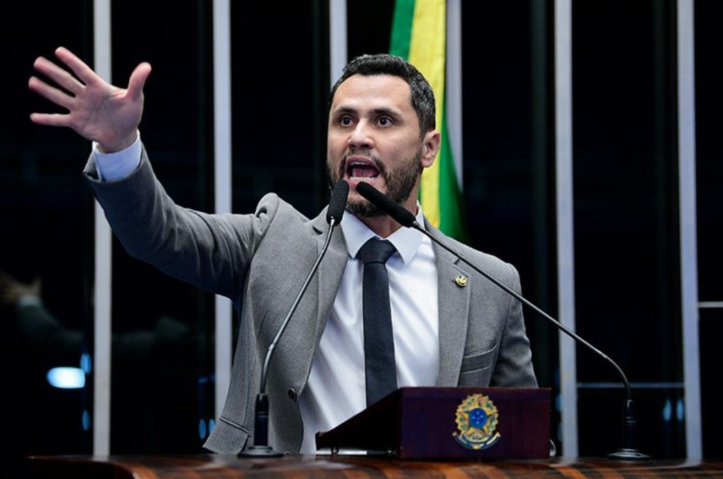 Senador Cleitinho Azevedo, de terno cinza e gravata preta, gesticula com a mão direita levantada enquanto fala com expressão firme ao microfone no plenário do Senado. Ao fundo, vê-se parte da bandeira do Brasil e o painel característico da Casa Legislativa.