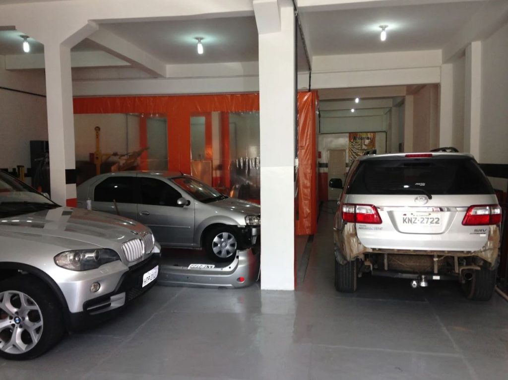 Funcionário da oficina Só Retoques manuseando a divisória de uma cabine de pintura, com veículos em processo de reparo automotivo.