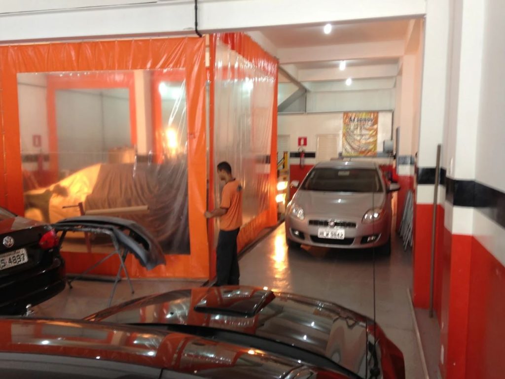 Funcionário da oficina Só Retoques manuseando a divisória de uma cabine de pintura, com veículos em processo de reparo automotivo.