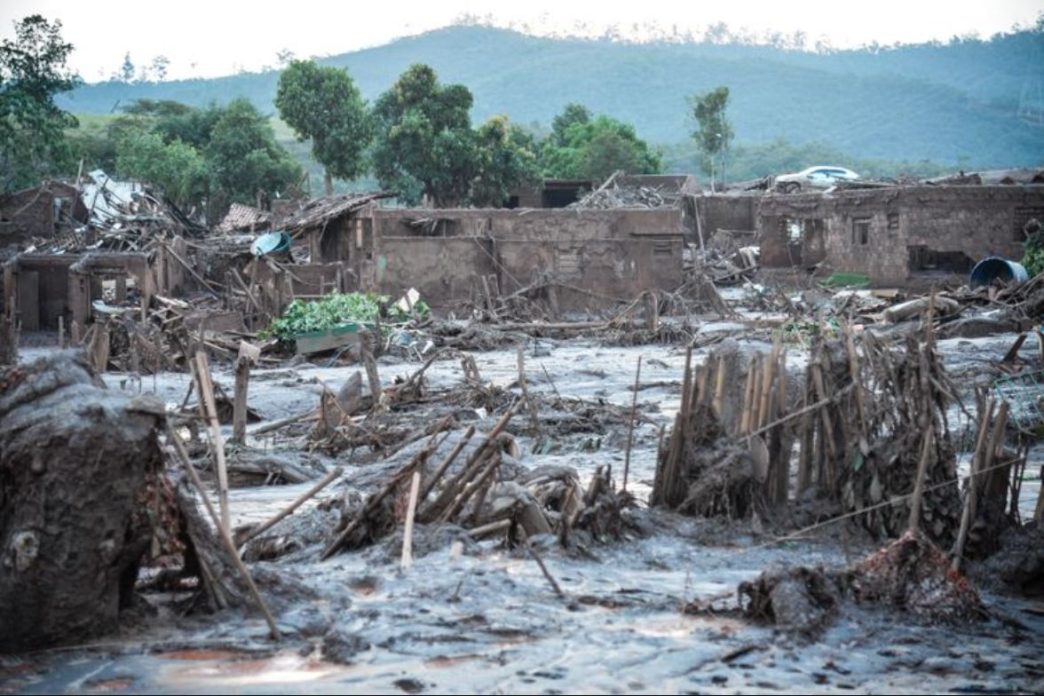 Área completamente destruída pela lama tóxica do rompimento da barragem de Fundão, em Mariana (MG), com casas soterradas e vegetação devastada.