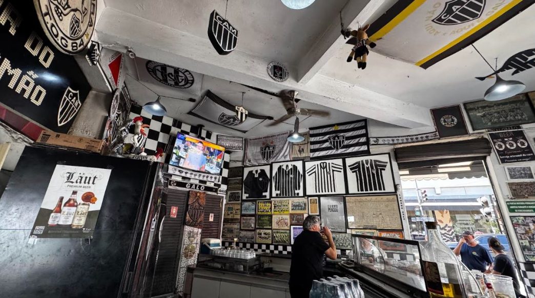 Interior do Bar do Salomão em Belo Horizonte, decorado com bandeiras, escudos e camisas do Clube Atlético Mineiro, com ambiente boêmio e elementos históricos nas paredes, incluindo televisão ligada, clientes ao balcão e iluminação tradicional.