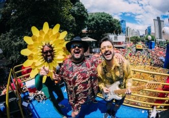 Três integrantes do bloco Beiço do Wando posam sorridentes sobre o trio elétrico durante apresentação no Carnaval de Belo Horizonte, com uma multidão colorida ao fundo. Um deles veste uma fantasia de flor amarela vibrante, outro traja roupa estampada com chapéu preto, e o terceiro segura uma guitarra elétrica vestindo camisa dourada translúcida. O céu está azul com algumas nuvens, e arranha-céus modernos compõem o cenário urbano.