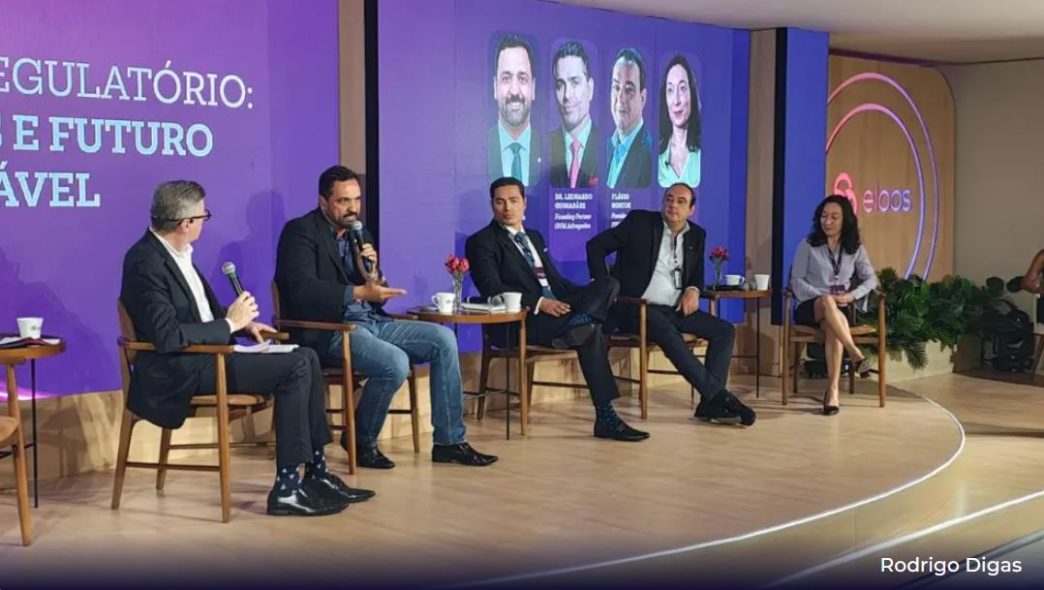 Painel de especialistas durante o primeiro Projeto Eloos, com cinco debatedores sentados no palco discutindo temas sobre regulação e sustentabilidade na mineração, em frente a um telão com os nomes dos participantes, no auditório da Mina de Águas Claras, em Nova Lima (MG).