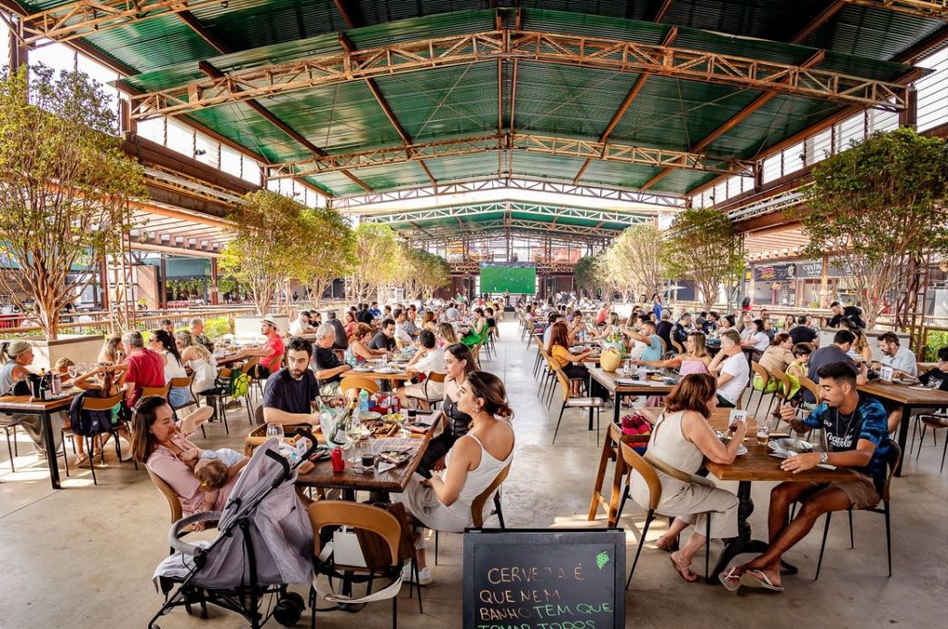 Público diverso aproveita o Festival de Pastel no Mercado de Origem, em Belo Horizonte, reunido em mesas sob galpão coberto e rodeado por árvores, em um ambiente arejado, familiar e gastronômico.