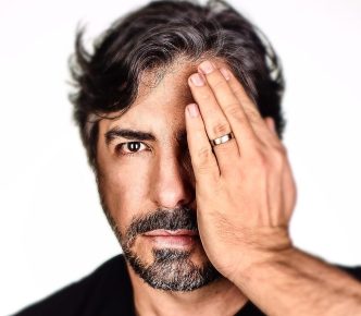 Retrato artístico de Fred Isak em fundo branco, cobrindo um dos olhos com a mão direita, com expressão séria e introspectiva.