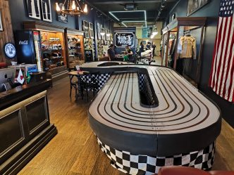 Pista de autorama montada no interior da barbearia Blackwood, em Belo Horizonte, com decoração retrô, bandeira dos EUA na parede e ambiente sofisticado, unindo entretenimento e estilo.