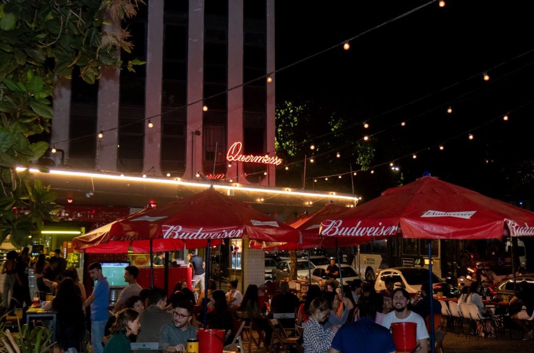 Clientes aproveitam o ambiente externo do bar Quermesse em Belo Horizonte à noite, sob luzes decorativas e guarda-sóis vermelhos da Budweiser, em clima descontraído na Praça Tiradentes.