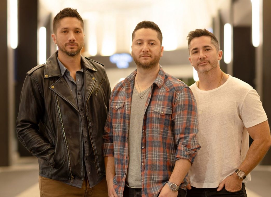Os irmãos Alejandro, Fabian e Daniel Manzano, da banda Boyce Avenue, posam lado a lado em ambiente interno iluminado, promovendo turnê no Brasil com show confirmado em Belo Horizonte.