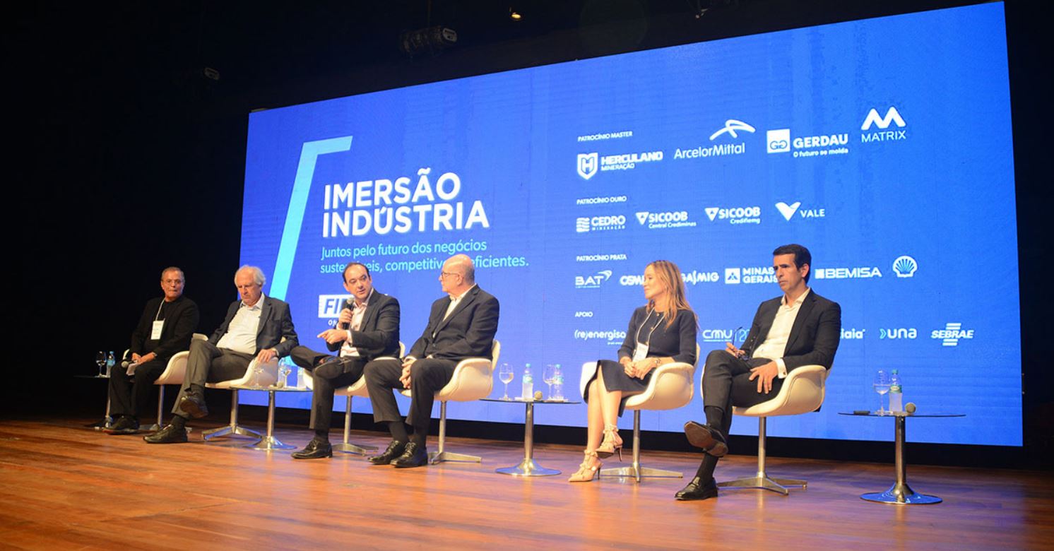 Painelistas participam de debate durante a edição anterior do Imersão Indústria, promovido pela FIEMG, em auditório com palco iluminado e tela azul exibindo logotipos de patrocinadores.