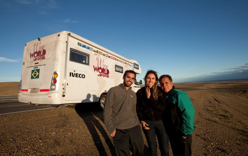 Três pessoas posam sorrindo ao lado de um motorhome branco com a logomarca “Wine World Adventure”, estacionado à beira de uma estrada deserta, sob céu azul. O veículo exibe bandeiras de diversos países e a bandeira do Brasil na traseira. Ao fundo, uma vasta paisagem árida se estende até o horizonte.
