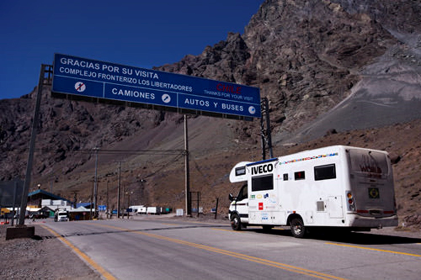 Motorhome do projeto Wine World Adventure cruza o complexo fronteiriço Los Libertadores, na Cordilheira dos Andes, entre Chile e Argentina. O veículo branco exibe bandeiras de diversos países e logos de patrocinadores, sob um céu azul intenso e rodeado por montanhas áridas e rochosas.