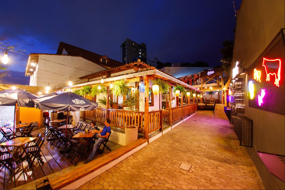 Fachada iluminada do restaurante Ô Chefe São Bento à noite, com varanda de madeira, plantas pendentes e mesas ao ar livre sob guarda-sóis.
