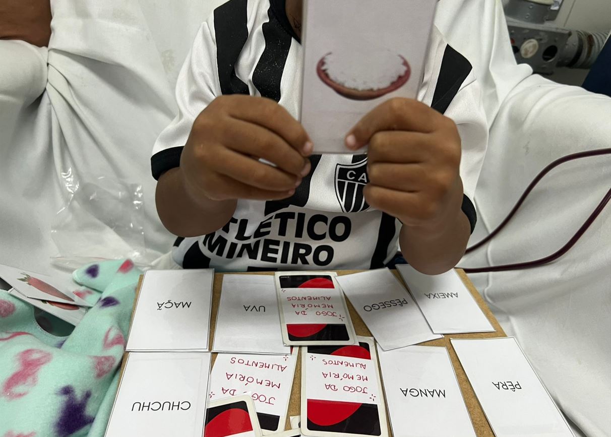 Criança em tratamento na Santa Casa BH segura carta educativa com imagem de alimento, durante atividade psicopedagógica no ambulatório.