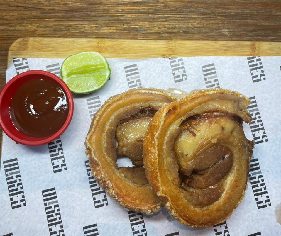 Porção de barriga de porco enrolada e crocante, servida com molho barbecue e limão, sobre papel personalizado do restaurante Ulisses.