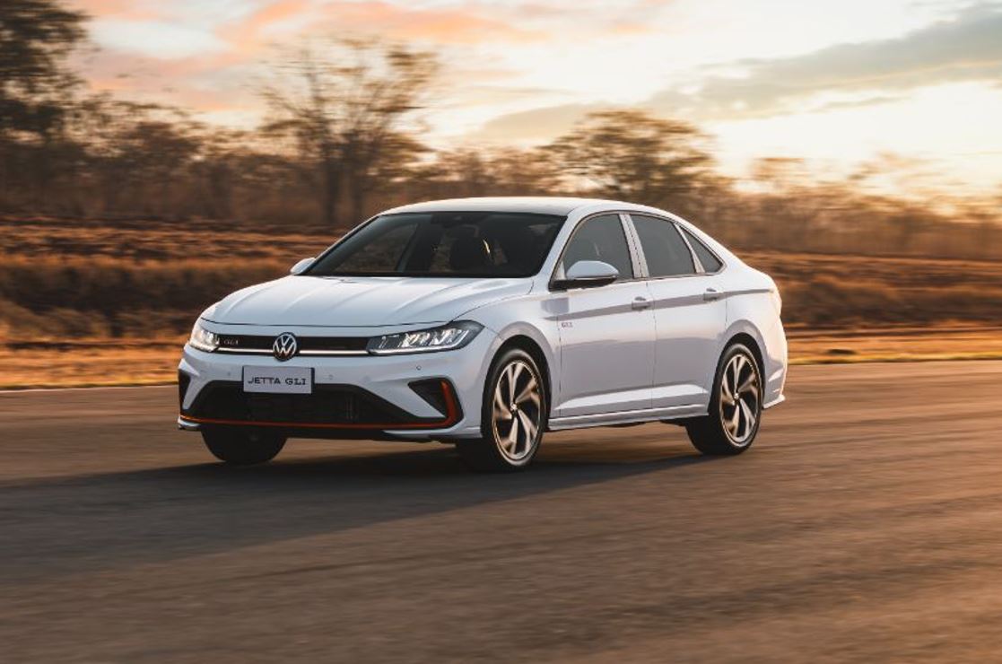 Volkswagen Jetta GLI 2026 na cor branco puro, em movimento em pista asfaltada ao entardecer, destacando design esportivo, grade colmeia com friso vermelho, rodas diamantadas aro 18 e assinatura visual em LED.